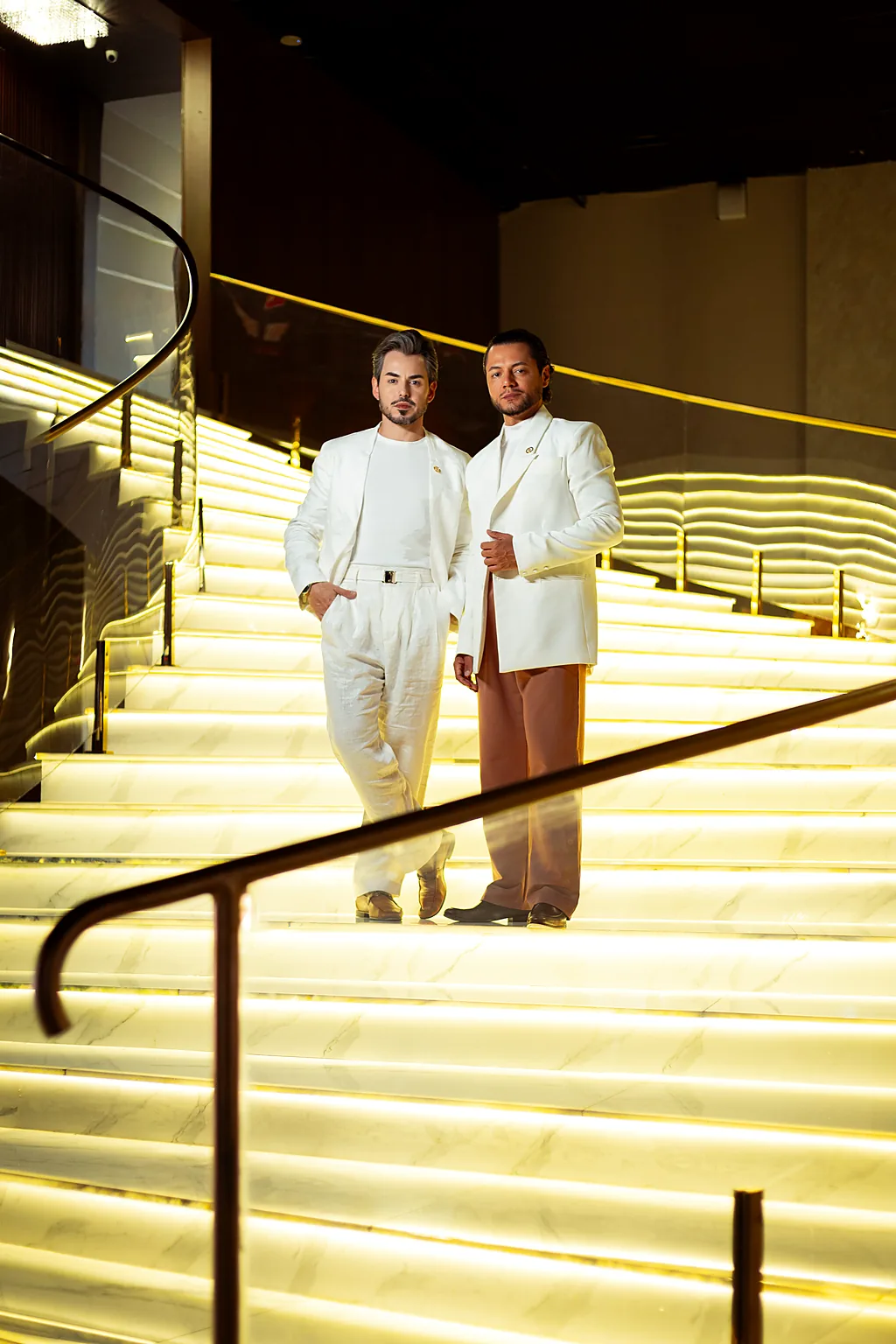 Maykon e Gleison, fundadores da High Beauty, lado a lado em trajes claros numa escadaria de mármore iluminada em dourado